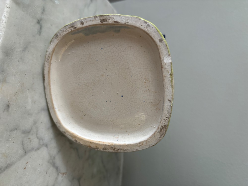 A small ceramic cottage-shaped money box featuring a blue thatched roof, white brick walls decorated with painted floral sprigs, and a brown arched entrance. View V