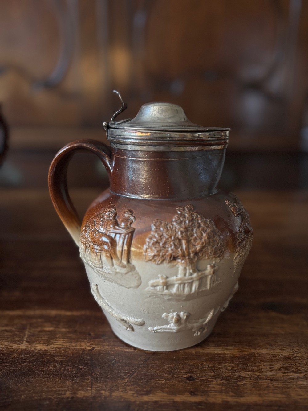An 18th-century Mortlake salt-glazed stoneware ale pitcher with raised tavern sprigged reliefs and a silver-plated hinged lid.