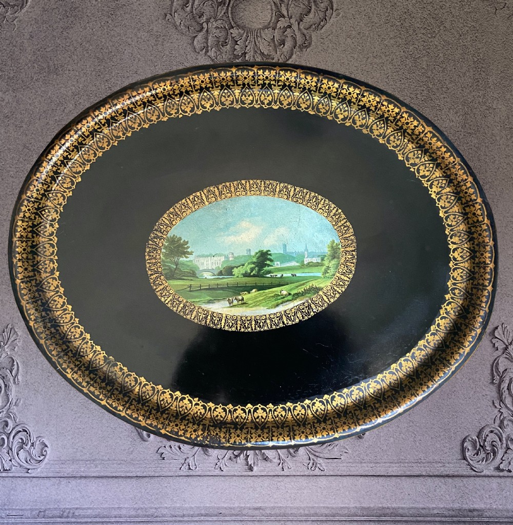 An oval black tole ware tray featuring intricate gold gilded borders around a central painted pastoral landscape scene.