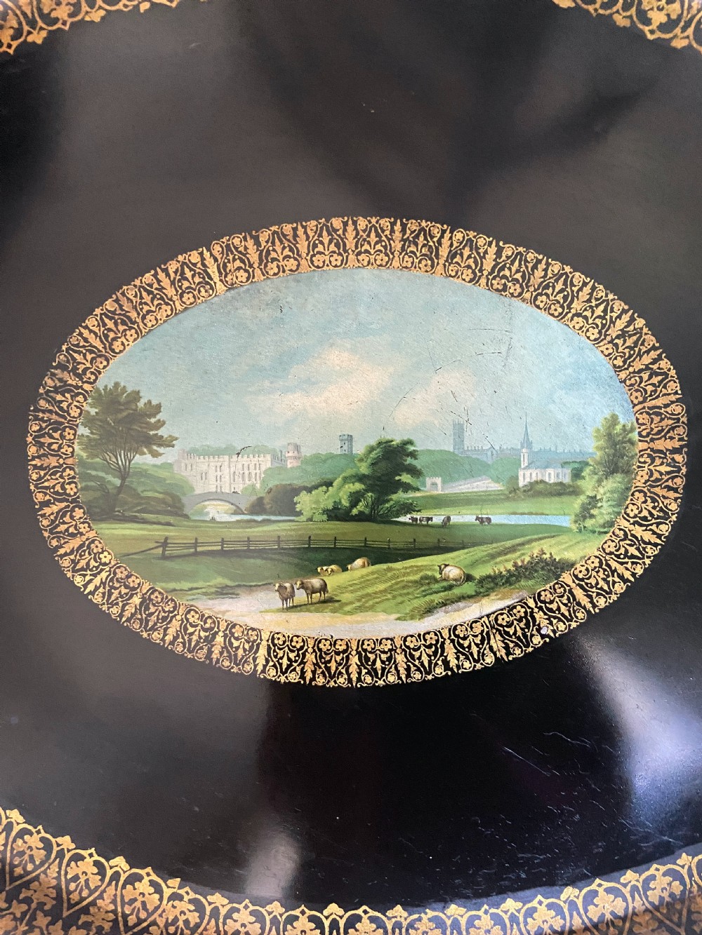 An oval black tole ware tray featuring intricate gold gilded borders around a central painted pastoral landscape scene. View IV