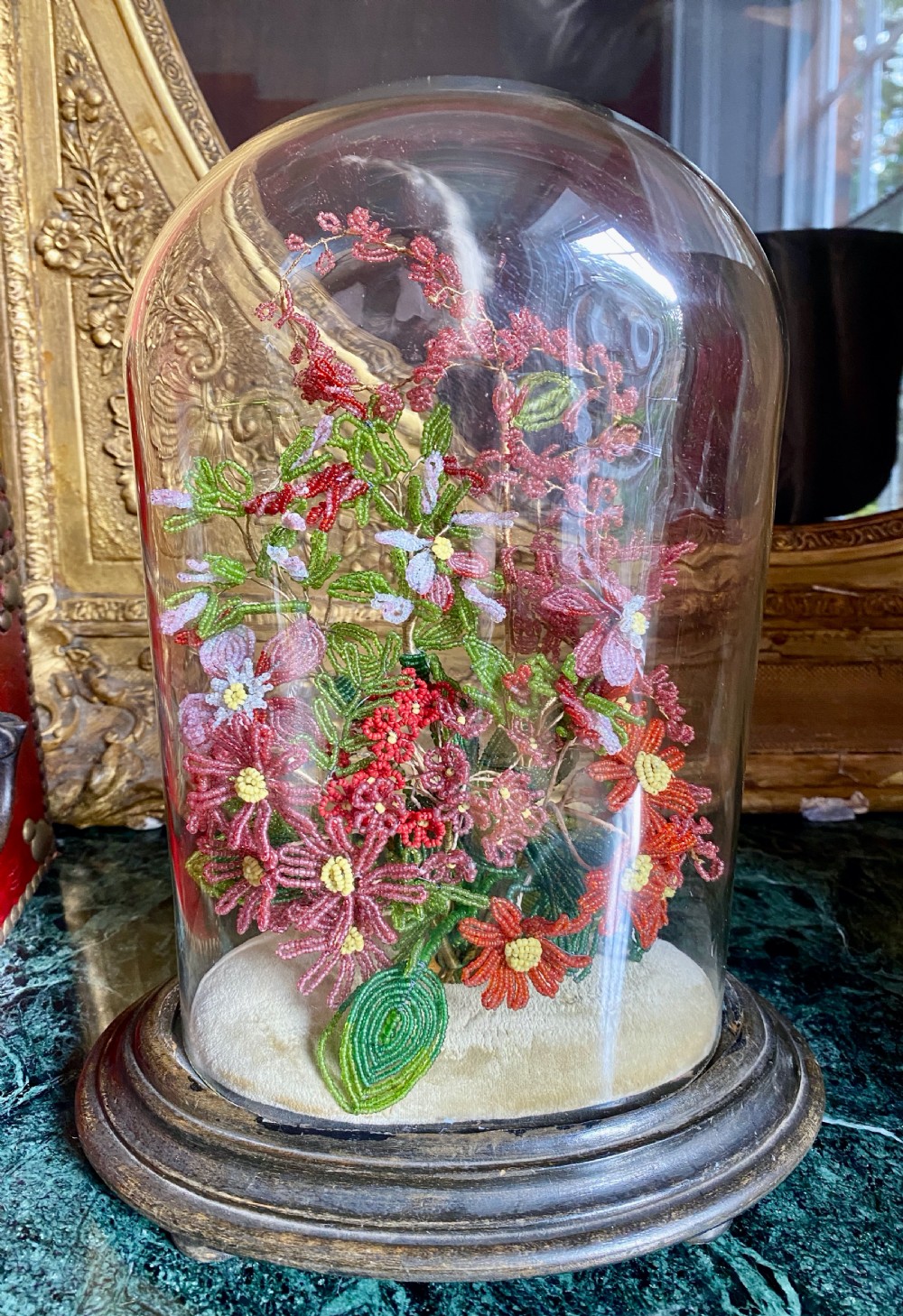 A clear glass parlour dome on a turned dark wooden base enclosing a French beaded floral bouquet with red, pink, and green seed beads.