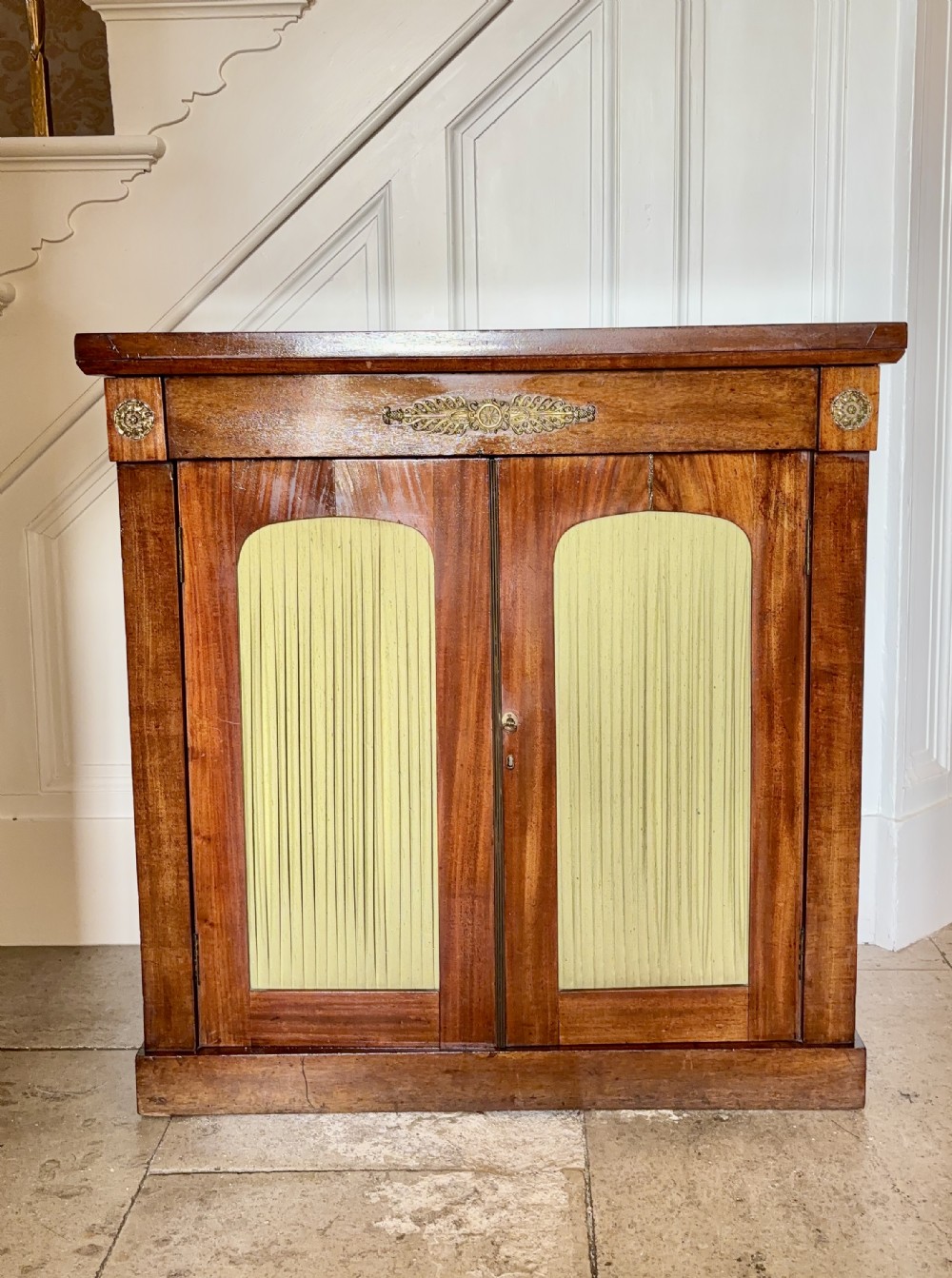 A late Regency rosewood side cabinet featuring two doors with arched pleated fabric panels and central brass decorative mounts.