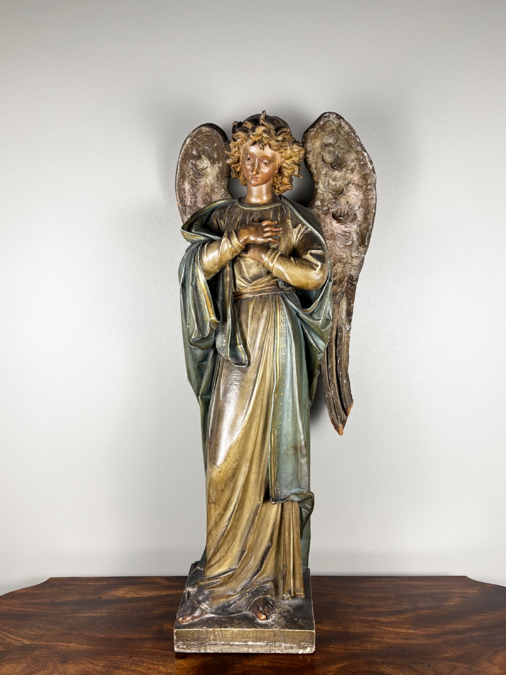 A 19th-century Italian carved wooden angel sculpture featuring extended feathered wings, muted green draped robes, and crossed hands over the chest on a square base.