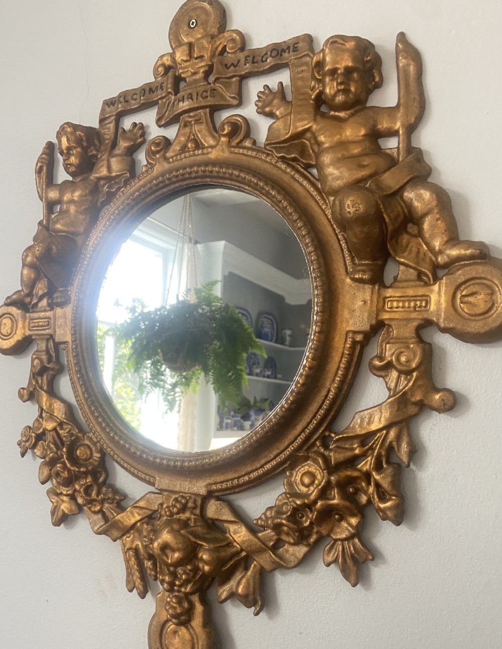 A circular cast iron wall mirror featuring a gold finish, flanked by sculpted cherubs holding a banner inscribed with Welcome Thrice Welcome. View III