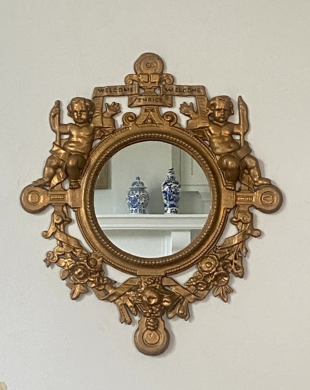 A circular cast iron wall mirror featuring a gold finish, flanked by sculpted cherubs holding a banner inscribed with Welcome Thrice Welcome.