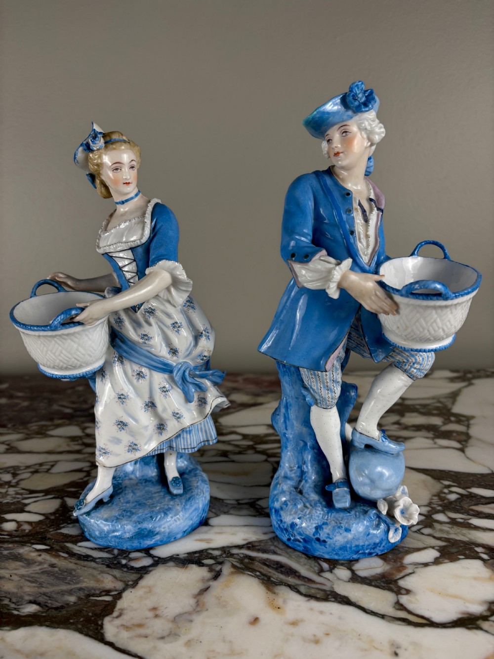 A pair of 19th-century French glazed porcelain figures in blue and white Rococo costume, each holding a woven basket.