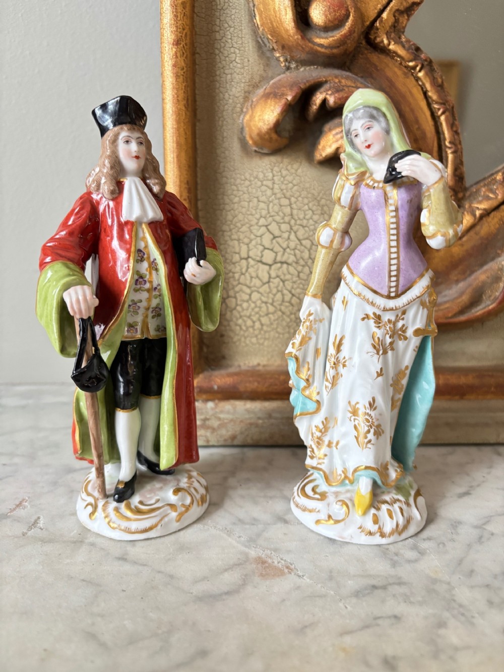A pair of hand-painted French porcelain figurines depicting an 18th-century man and woman in period costume standing on gilded scroll bases.
