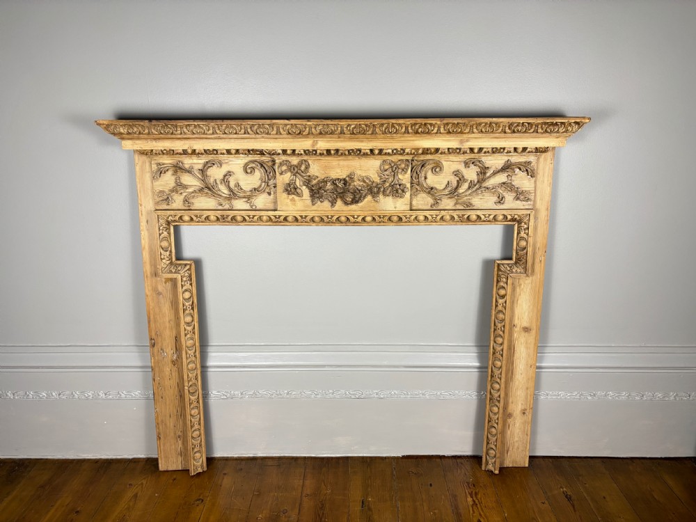 A carved Georgian pine fire surround featuring egg-and-dart moulding, floral swags, and ribbon motifs in a natural timber finish.