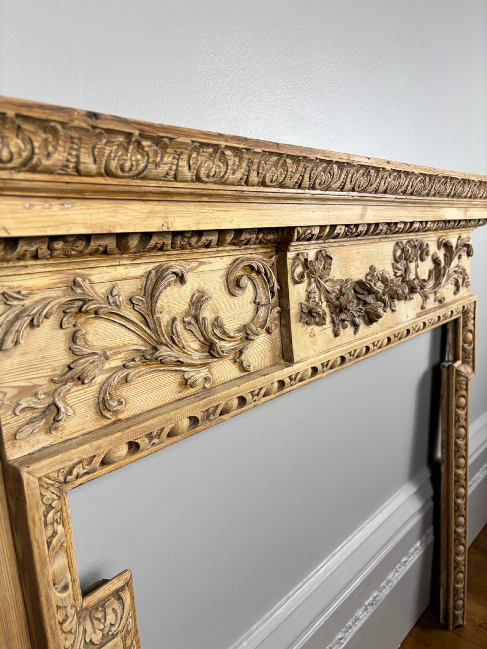 A carved Georgian pine fire surround featuring egg-and-dart moulding, floral swags, and ribbon motifs in a natural timber finish. View III