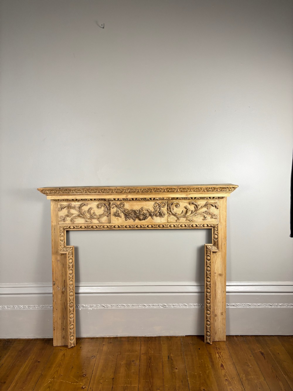 A carved Georgian pine fire surround featuring egg-and-dart moulding, floral swags, and ribbon motifs in a natural timber finish. View V