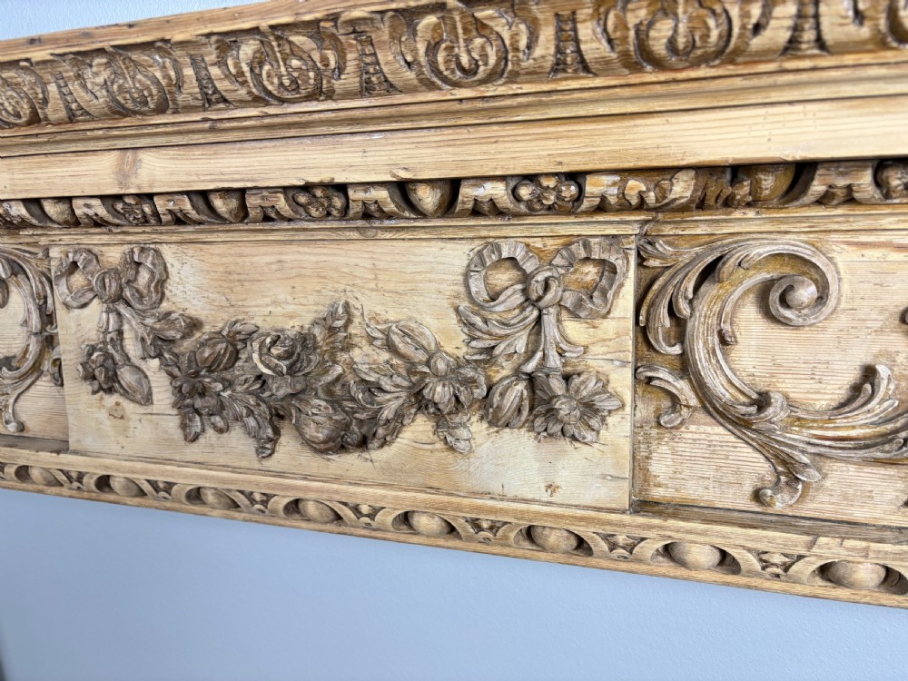 A carved Georgian pine fire surround featuring egg-and-dart moulding, floral swags, and ribbon motifs in a natural timber finish. View VI