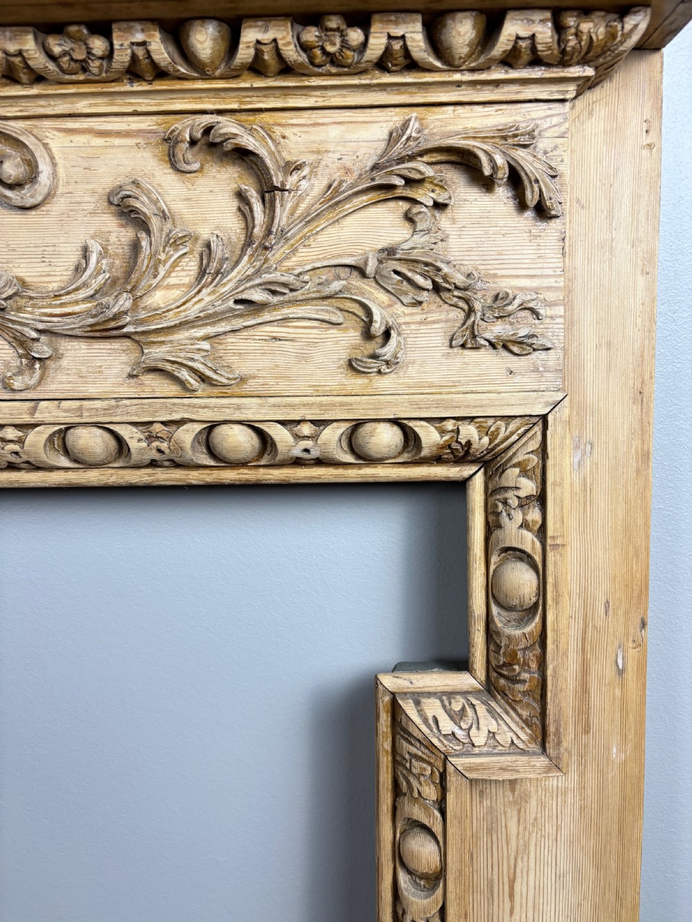 A carved Georgian pine fire surround featuring egg-and-dart moulding, floral swags, and ribbon motifs in a natural timber finish. View VII