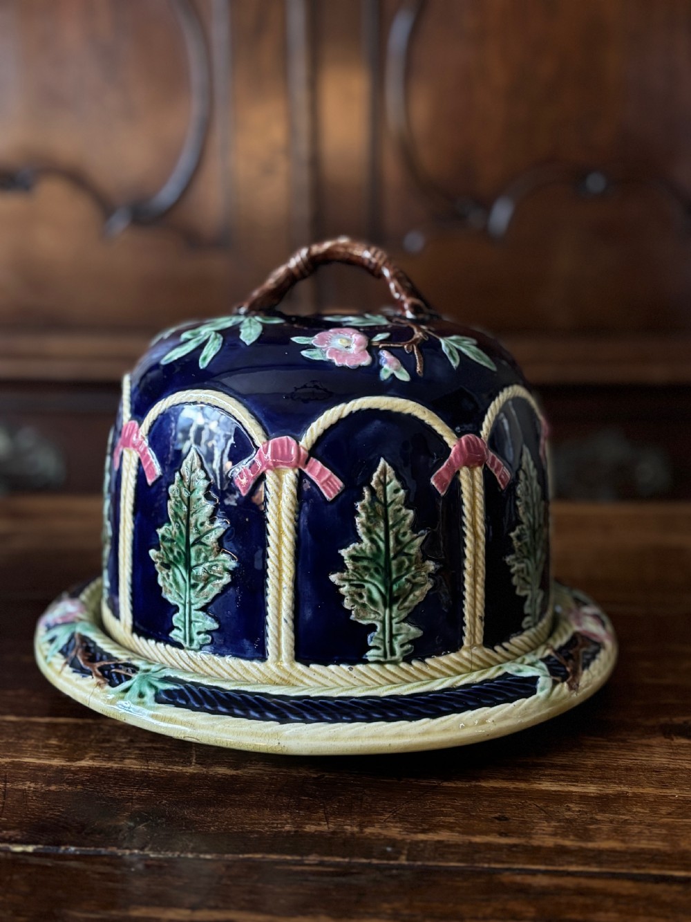 A Victorian majolica cheese dome featuring a cobalt blue glazed body, relief-moulded green leaves, cream rope arches, and pink bows, set on a matching round base.