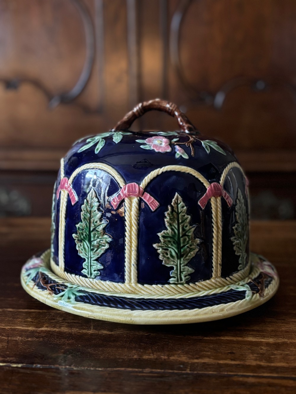 A Victorian majolica cheese dome featuring a cobalt blue glazed body, relief-moulded green leaves, cream rope arches, and pink bows, set on a matching round base. View II