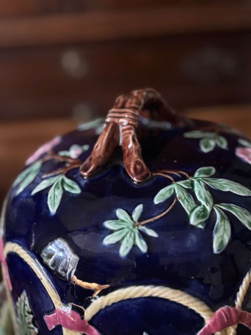 A Victorian majolica cheese dome featuring a cobalt blue glazed body, relief-moulded green leaves, cream rope arches, and pink bows, set on a matching round base. View III