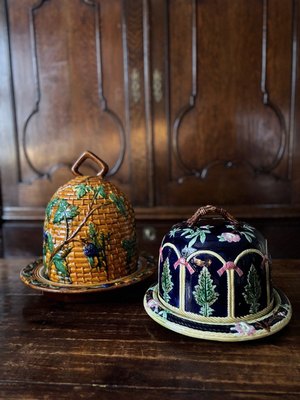 A Victorian majolica cheese dome featuring a cobalt blue glazed body, relief-moulded green leaves, cream rope arches, and pink bows, set on a matching round base. View VIII