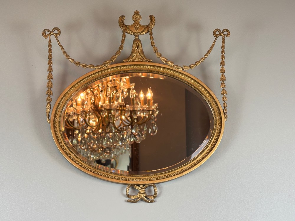 An oval gold giltwood wall mirror featuring a top urn cresting, ribbon bow accents, draped husk garlands, and an inner beaded rim. View VI