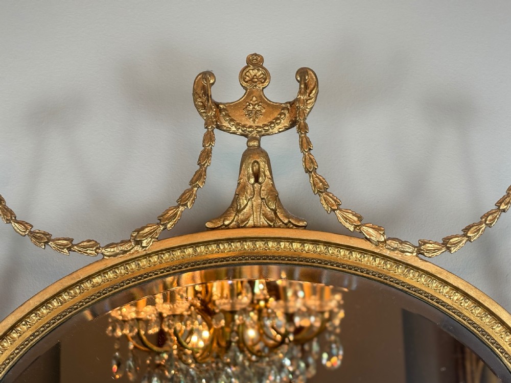 An oval gold giltwood wall mirror featuring a top urn cresting, ribbon bow accents, draped husk garlands, and an inner beaded rim. View II