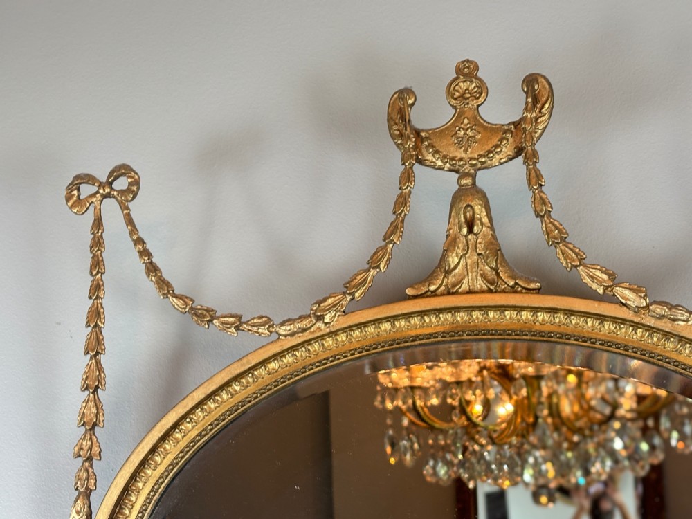 An oval gold giltwood wall mirror featuring a top urn cresting, ribbon bow accents, draped husk garlands, and an inner beaded rim. View III
