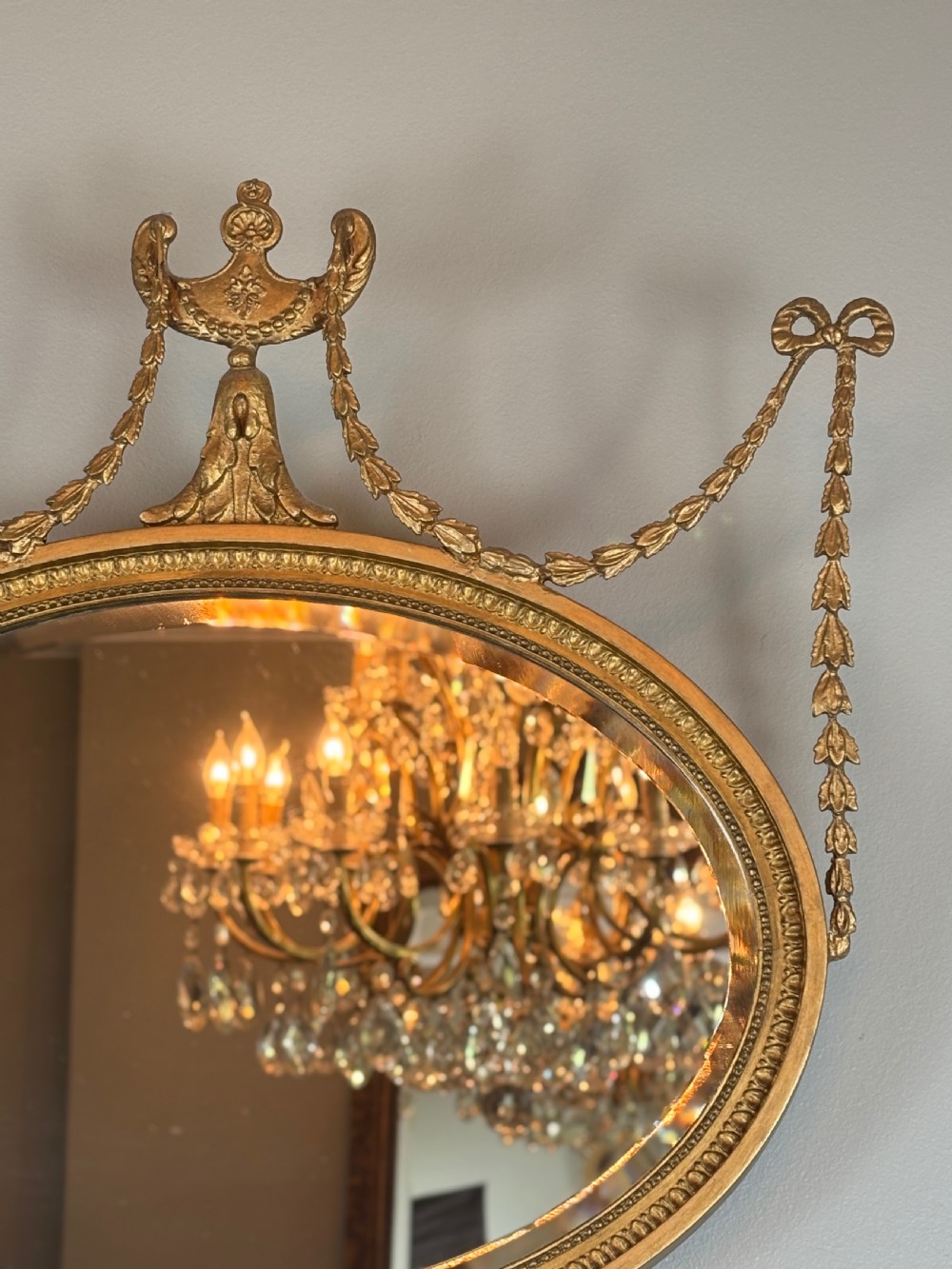 An oval gold giltwood wall mirror featuring a top urn cresting, ribbon bow accents, draped husk garlands, and an inner beaded rim. View V