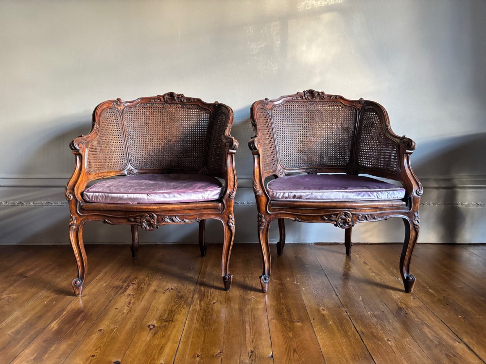 A pair of Louis XV style carved wooden bergere armchairs featuring woven cane backs and purple velvet seat cushions.