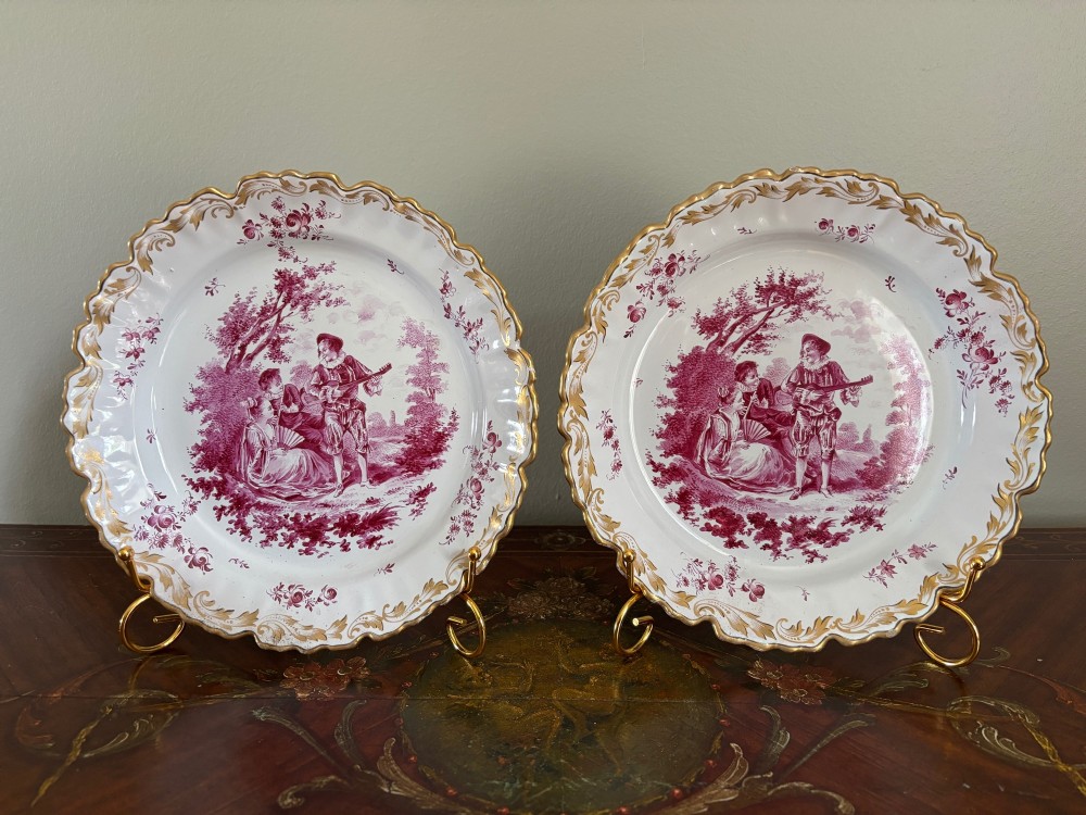 A pair of circular tin-glazed faience dishes featuring central pastoral figures rendered in magenta puce camaieu with gilded scalloped edges.