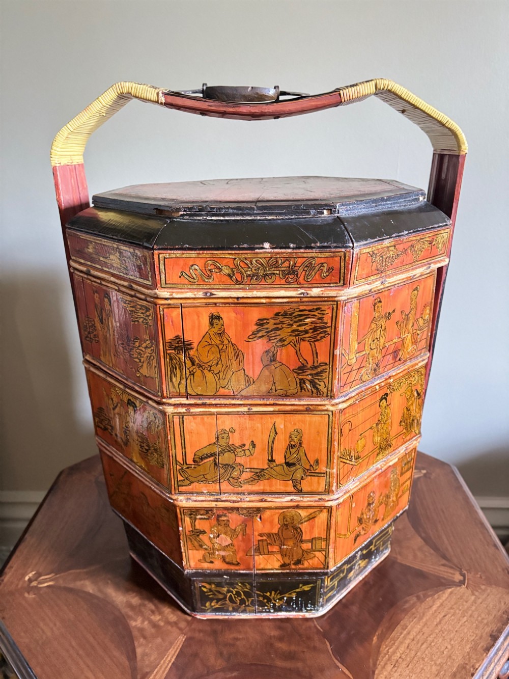 An octagonal red and black lacquered three-tier Chinese dowry basket with an overhead bentwood handle bound in woven cane and panels painted with figures.