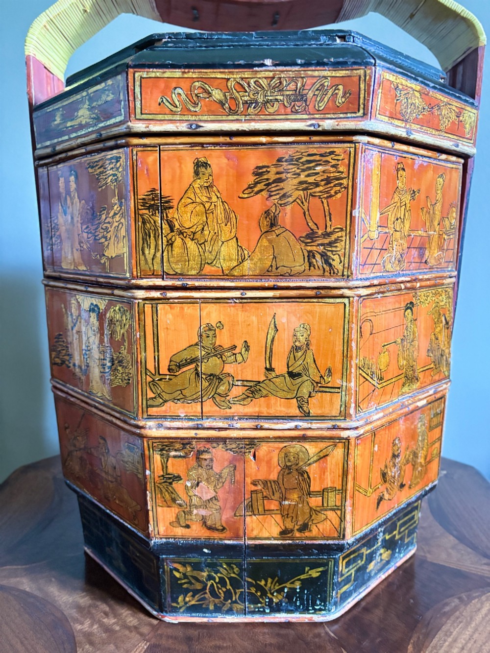 An octagonal red and black lacquered three-tier Chinese dowry basket with an overhead bentwood handle bound in woven cane and panels painted with figures. View II
