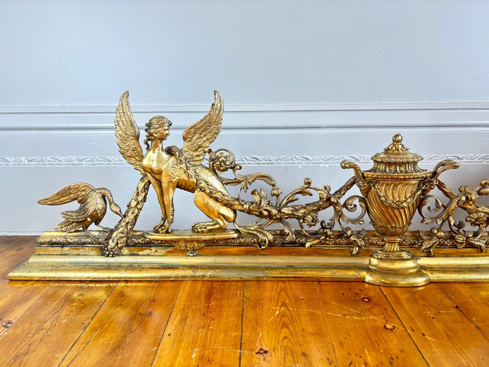 A long gilt bronze fireplace fender featuring winged sphinxes on either side, foliate scrollwork, and a central classical urn on a stepped base. View II