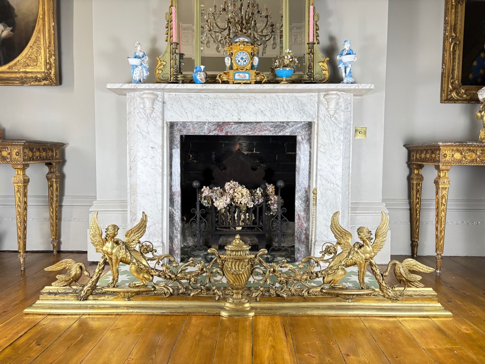 A long gilt bronze fireplace fender featuring winged sphinxes on either side, foliate scrollwork, and a central classical urn on a stepped base. View IX