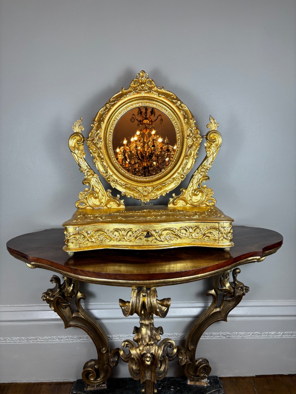 An 18th-century Italian giltwood table mirror featuring an oval glass frame supported by ornate scroll carved arms atop a single-drawer plinth base. View II