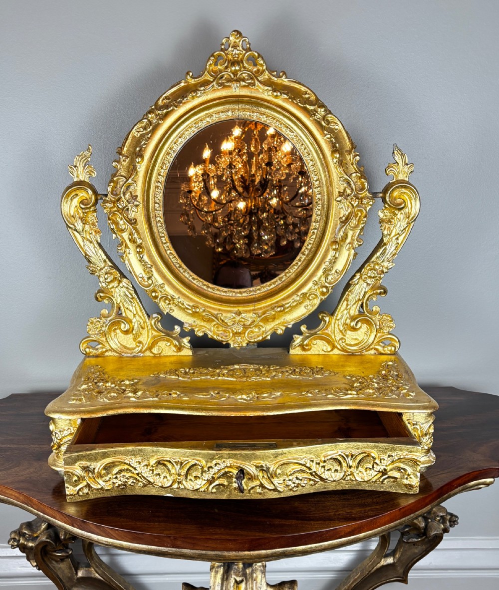 An 18th-century Italian giltwood table mirror featuring an oval glass frame supported by ornate scroll carved arms atop a single-drawer plinth base. View IV