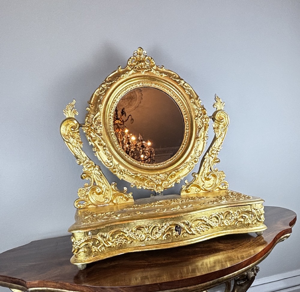An 18th-century Italian giltwood table mirror featuring an oval glass frame supported by ornate scroll carved arms atop a single-drawer plinth base. View V