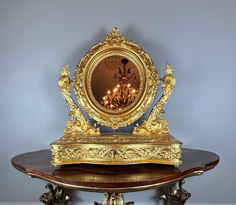 An 18th-century Italian giltwood table mirror featuring an oval glass frame supported by ornate scroll carved arms atop a single-drawer plinth base.