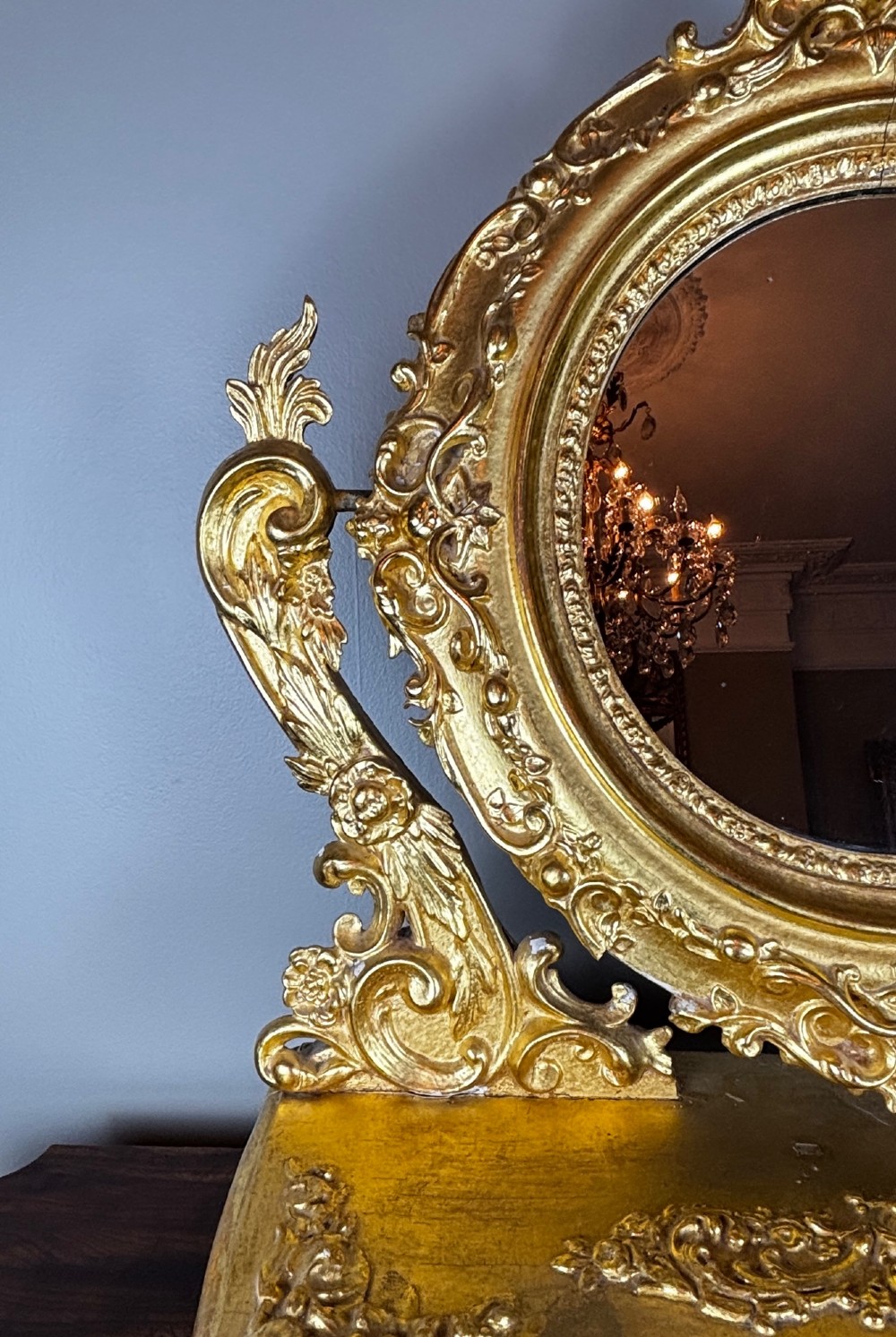 An 18th-century Italian giltwood table mirror featuring an oval glass frame supported by ornate scroll carved arms atop a single-drawer plinth base. View VI