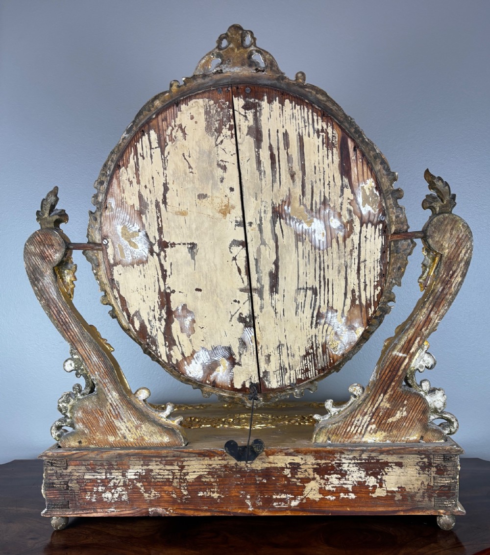 An 18th-century Italian giltwood table mirror featuring an oval glass frame supported by ornate scroll carved arms atop a single-drawer plinth base. View VIII