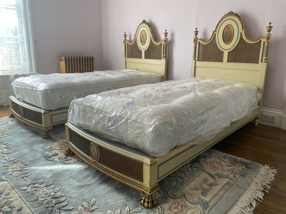 A pair of antique cream and gilt painted single beds featuring caned headboards with central painted portrait medallions and carved paw feet.