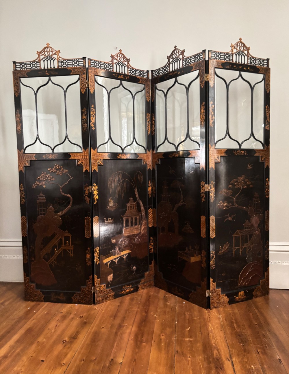 A four-fold black lacquered screen with Chinese Chippendale fretwork crestings, glazed upper panels, and incised gold chinoiserie landscape scenes.