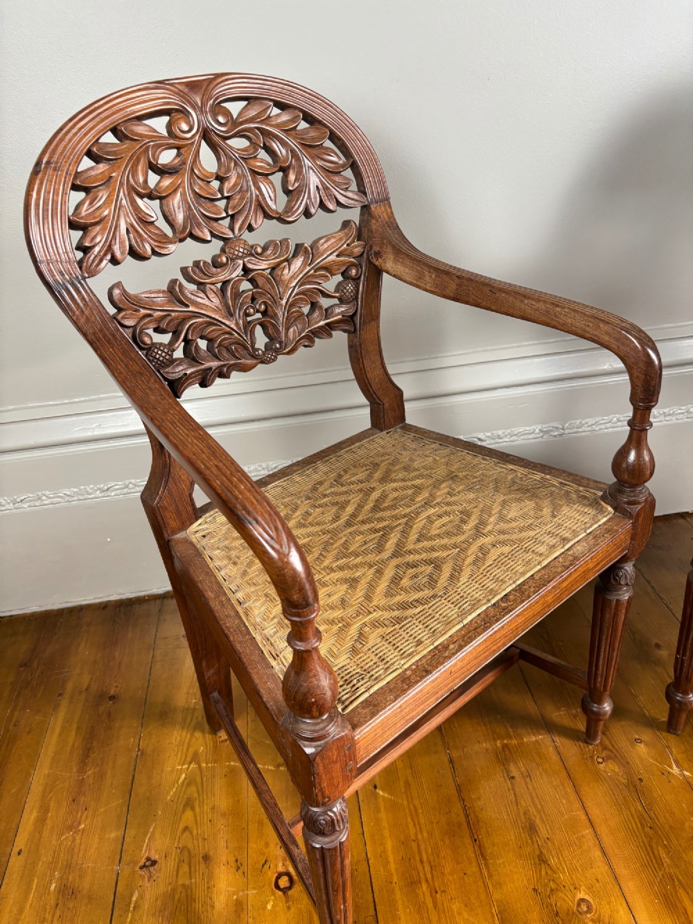 A pair of carved hardwood open armchairs featuring intricate openwork foliate backrests, swept arms, woven cane seats, and turned front legs. View II