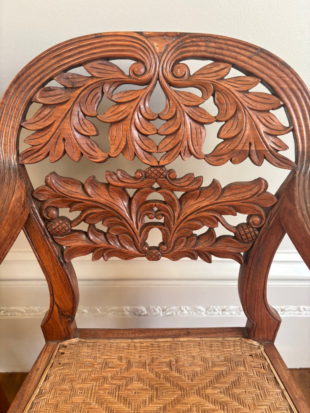 A pair of carved hardwood open armchairs featuring intricate openwork foliate backrests, swept arms, woven cane seats, and turned front legs. View V