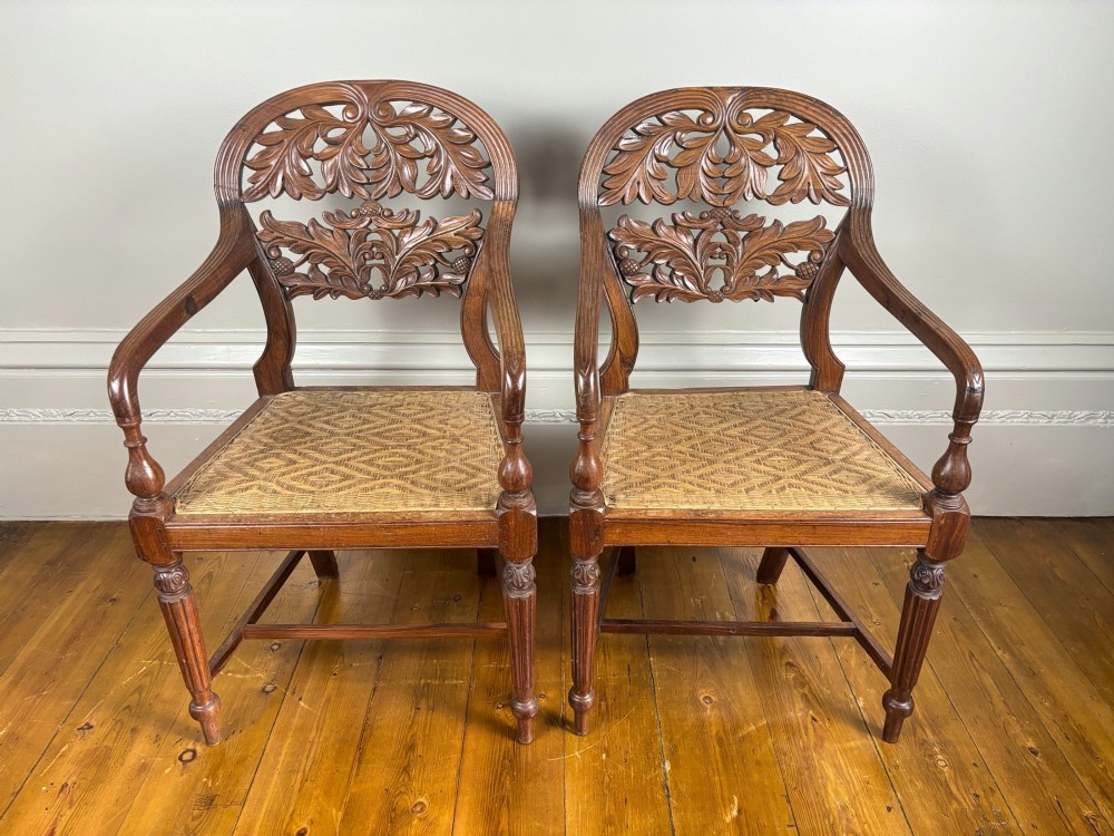 A pair of carved hardwood open armchairs featuring intricate openwork foliate backrests, swept arms, woven cane seats, and turned front legs.