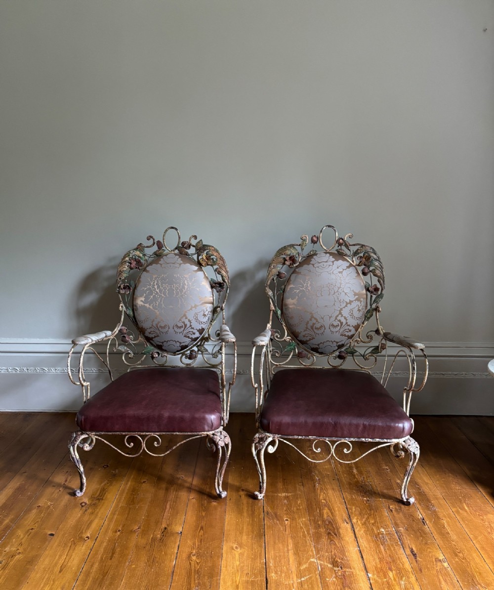 A pair of French wrought iron armchairs featuring cream painted scrolled frames, polychrome floral accents around oval backrests, and burgundy leather seats.