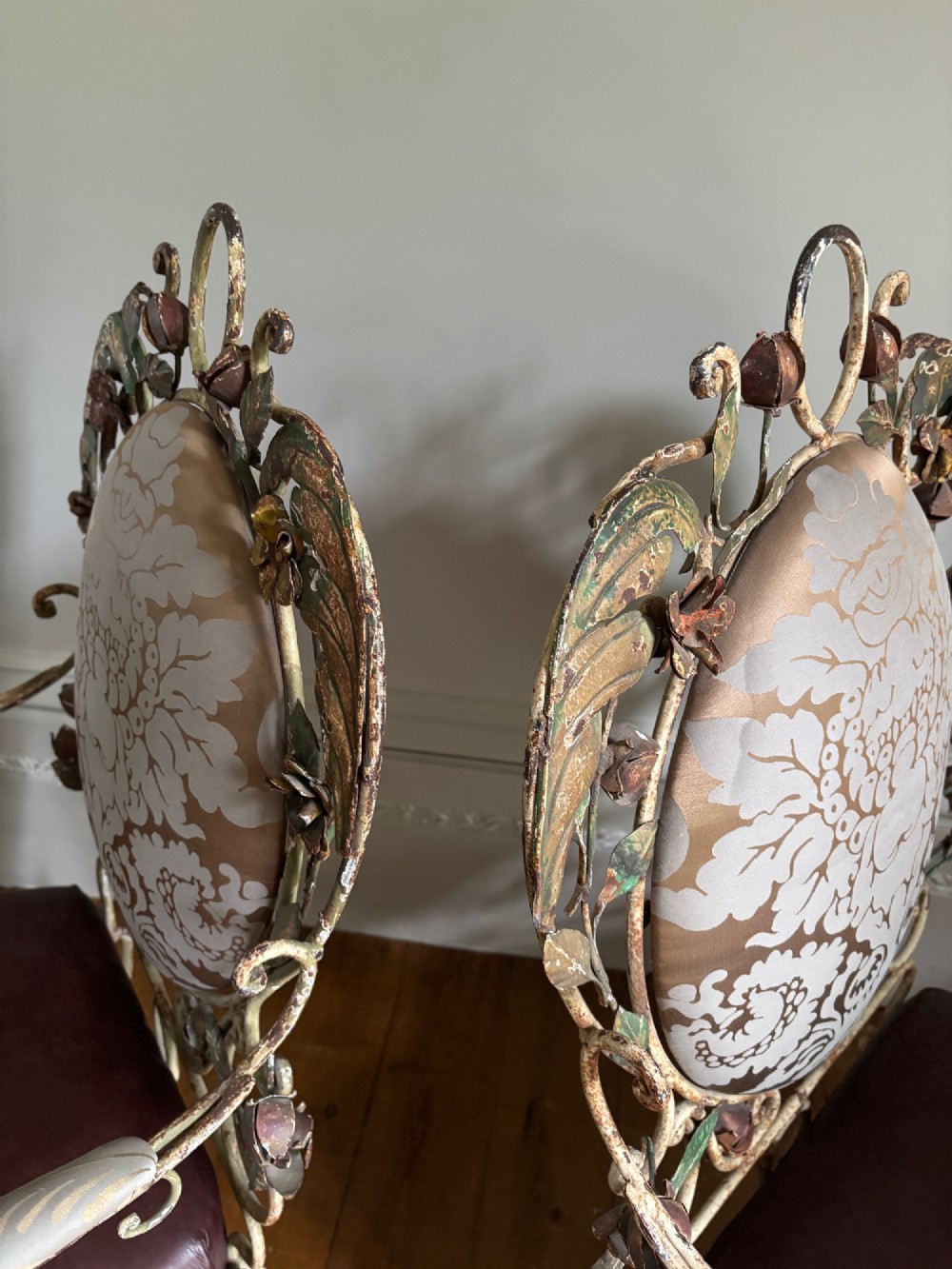 A pair of French wrought iron armchairs featuring cream painted scrolled frames, polychrome floral accents around oval backrests, and burgundy leather seats. View II