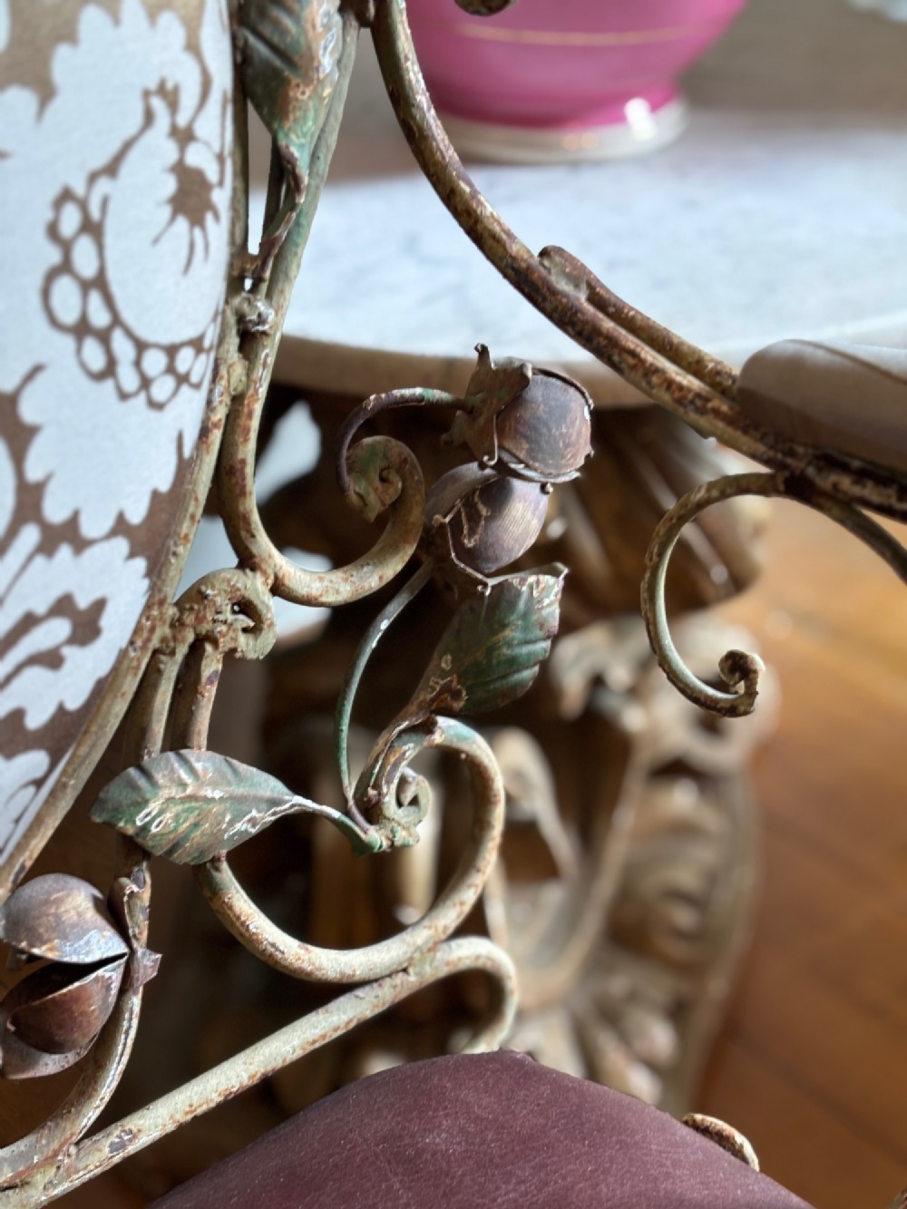 A pair of French wrought iron armchairs featuring cream painted scrolled frames, polychrome floral accents around oval backrests, and burgundy leather seats. View X