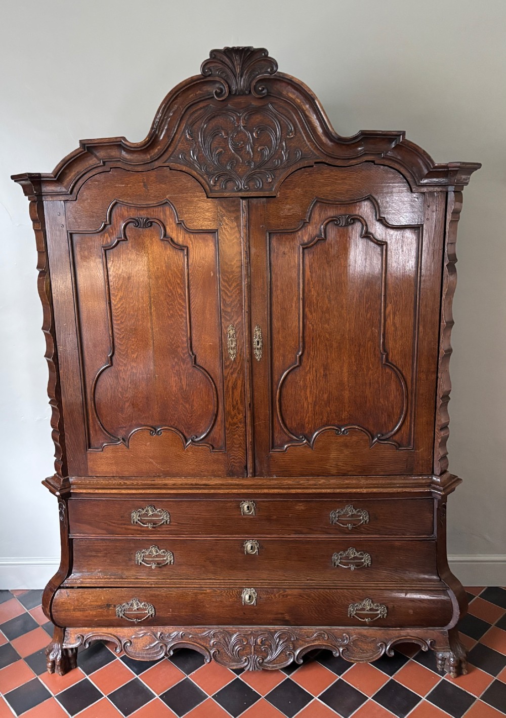 An 18th-century Dutch carved oak linen press featuring contoured panel doors, a carved cresting pediment, brass handles, and a bombee lower section with drawers.