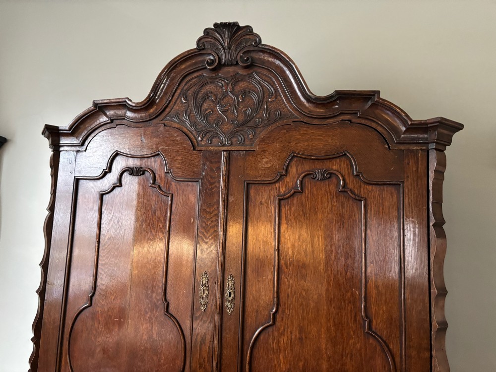 An 18th-century Dutch carved oak linen press featuring contoured panel doors, a carved cresting pediment, brass handles, and a bombee lower section with drawers. View III
