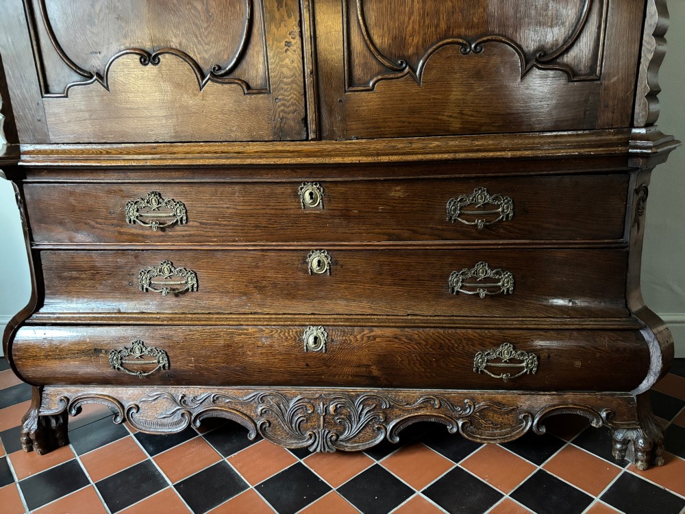 An 18th-century Dutch carved oak linen press featuring contoured panel doors, a carved cresting pediment, brass handles, and a bombee lower section with drawers. View IV