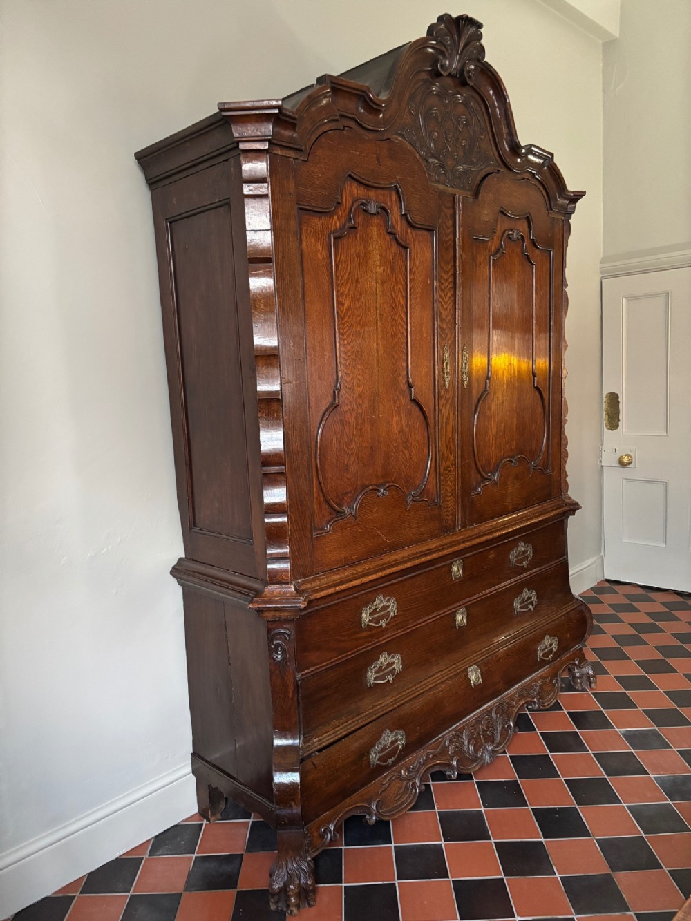An 18th-century Dutch carved oak linen press featuring contoured panel doors, a carved cresting pediment, brass handles, and a bombee lower section with drawers. View V