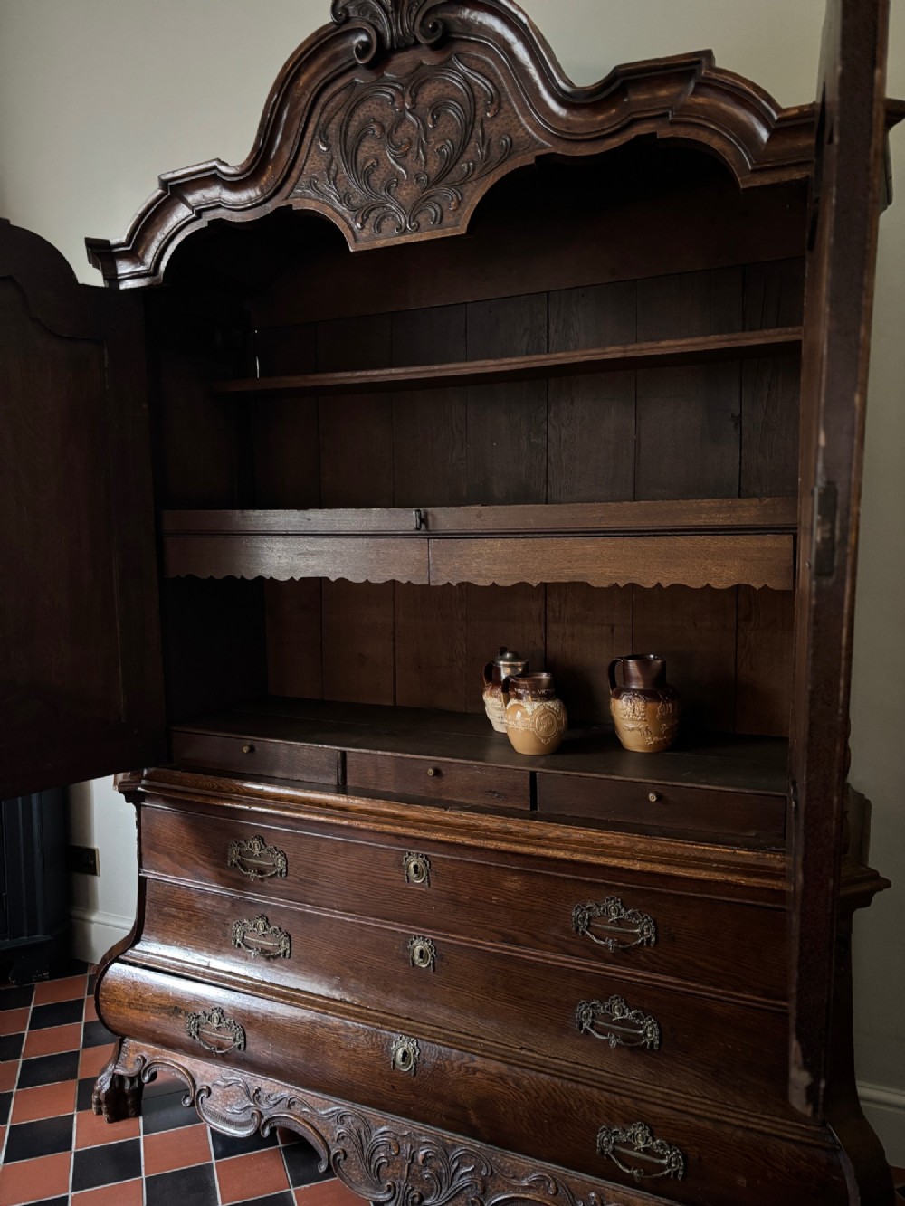 An 18th-century Dutch carved oak linen press featuring contoured panel doors, a carved cresting pediment, brass handles, and a bombee lower section with drawers. View VI