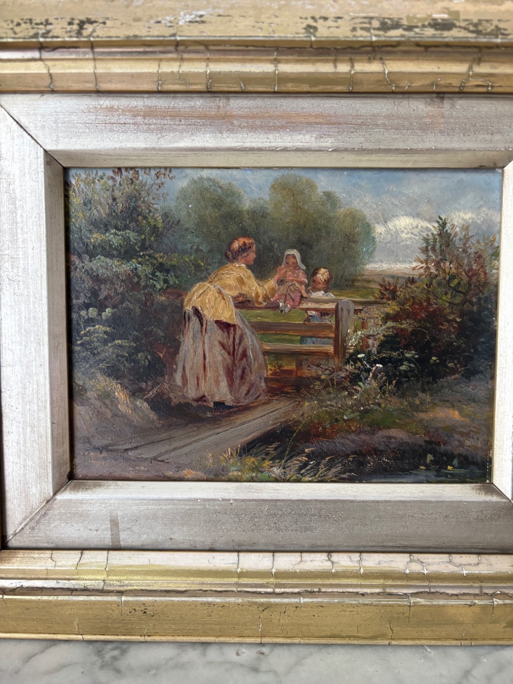 A small 19th-century style oil painting on board depicting a woman and children near a rustic wooden fence, set in a wide gilded frame. View VI